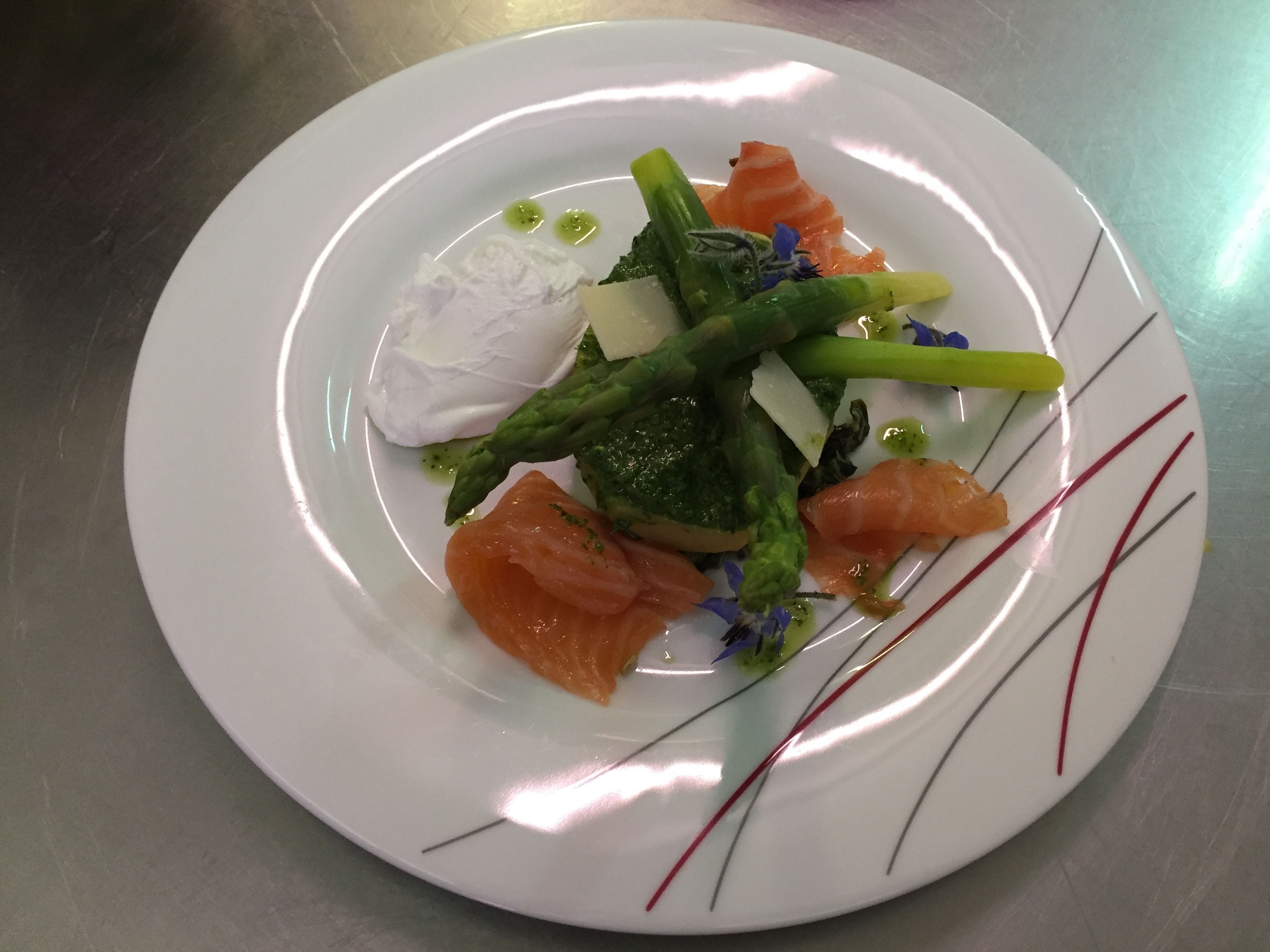 Un plato blanco con diversos alimentos, incluyendo salmón, espárragos y una cucharada de crema.