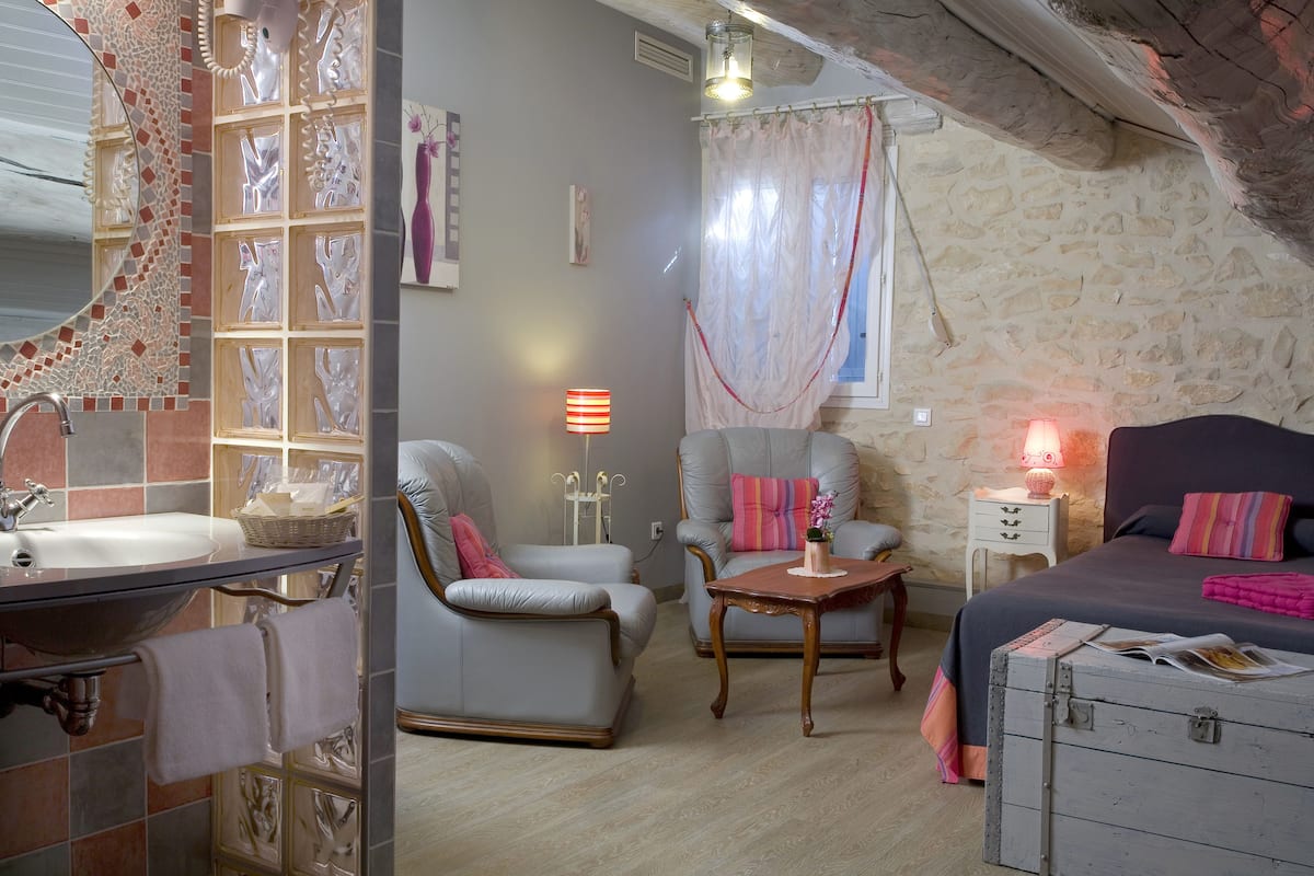 A cozy room with a bathroom, a bed, a chair, a table, and a dresser.