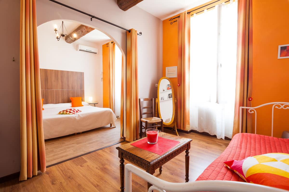 A hotel room with a bed, a small table with a drink, a chair, a mirror, and a window with curtains.