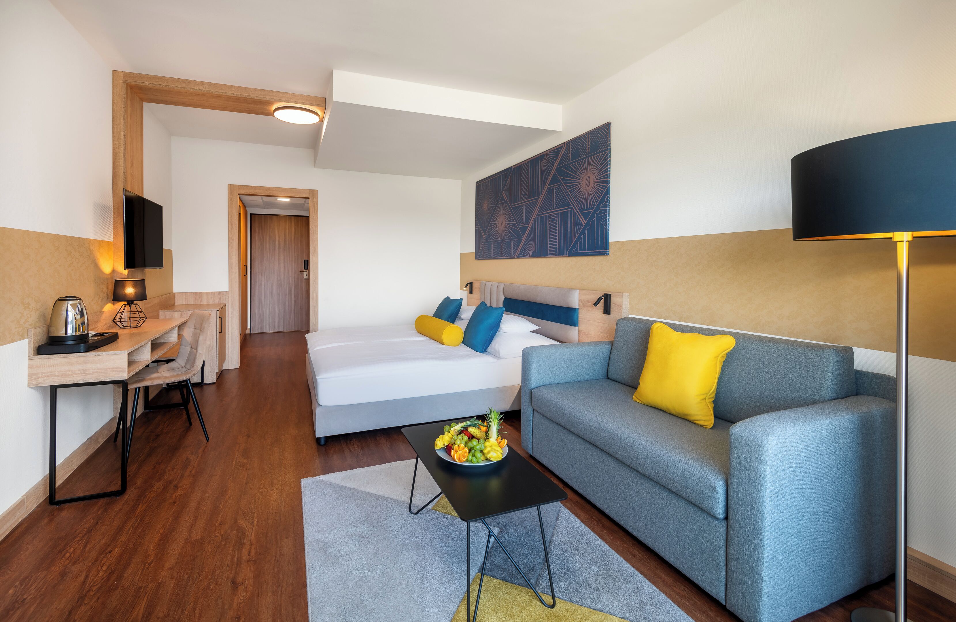 A modern hotel room with a sofa, a dining table, a flat-screen TV, and a large painting on the wall.
