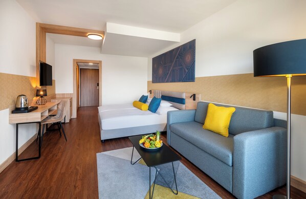 A modern hotel room with a sofa, a dining table, a flat-screen TV, and a large painting on the wall.