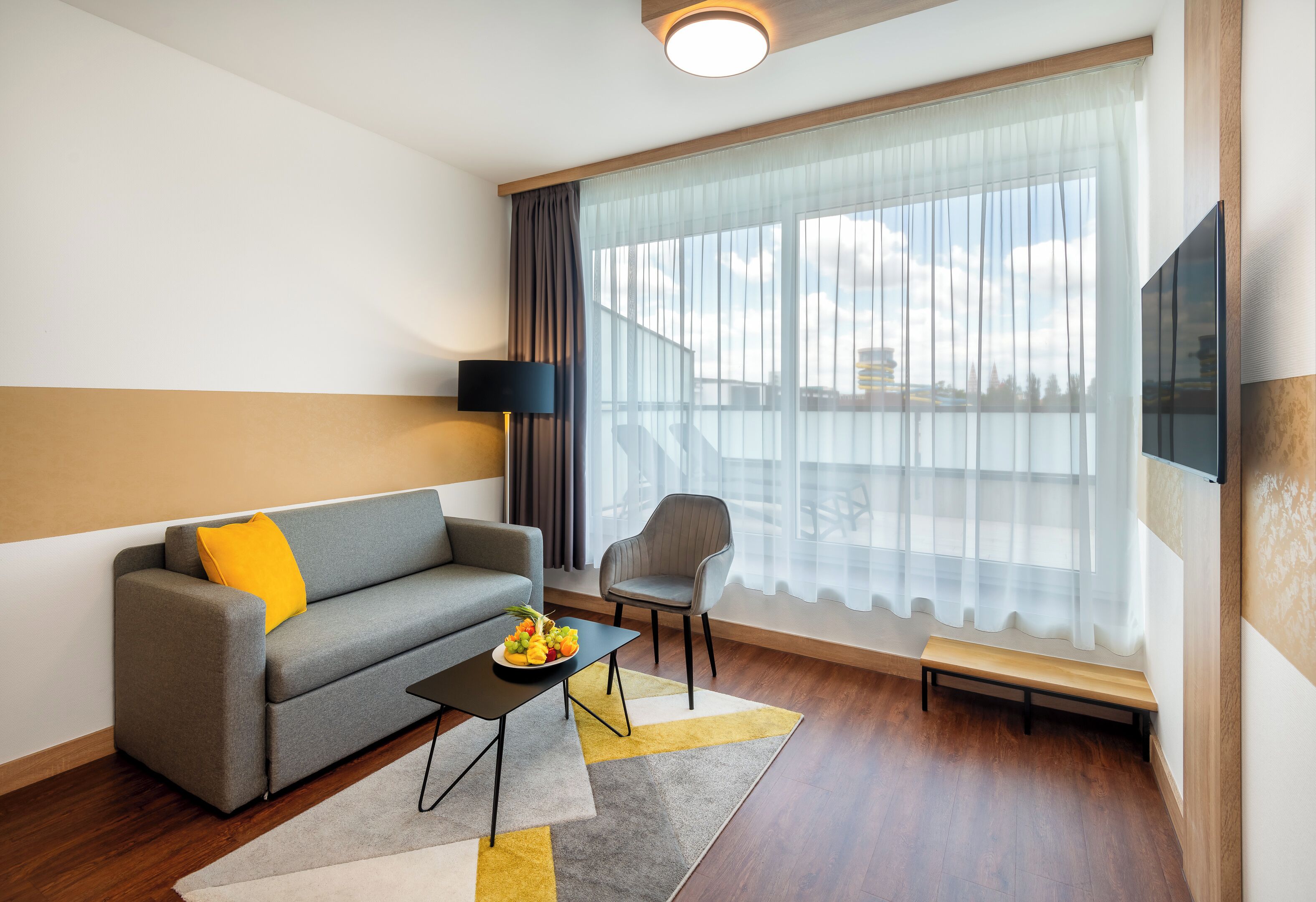 A modern living room with a grey sofa, a small table with fruit, a grey armchair, a bench, and a large window with curtains.