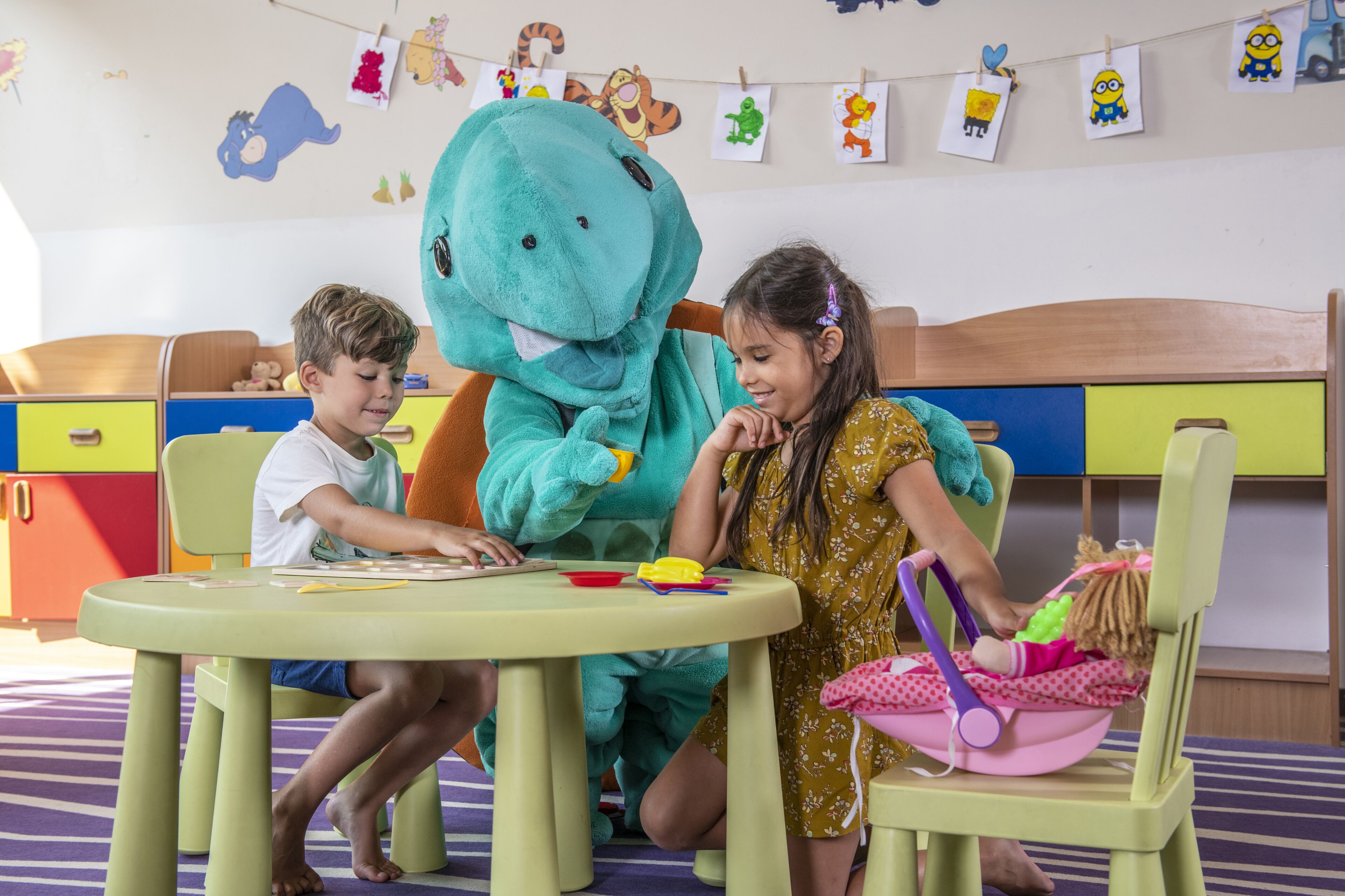 A child and an adult sitting at a table with a large plush toy in a room with colorful furniture and children's artwork on the walls.