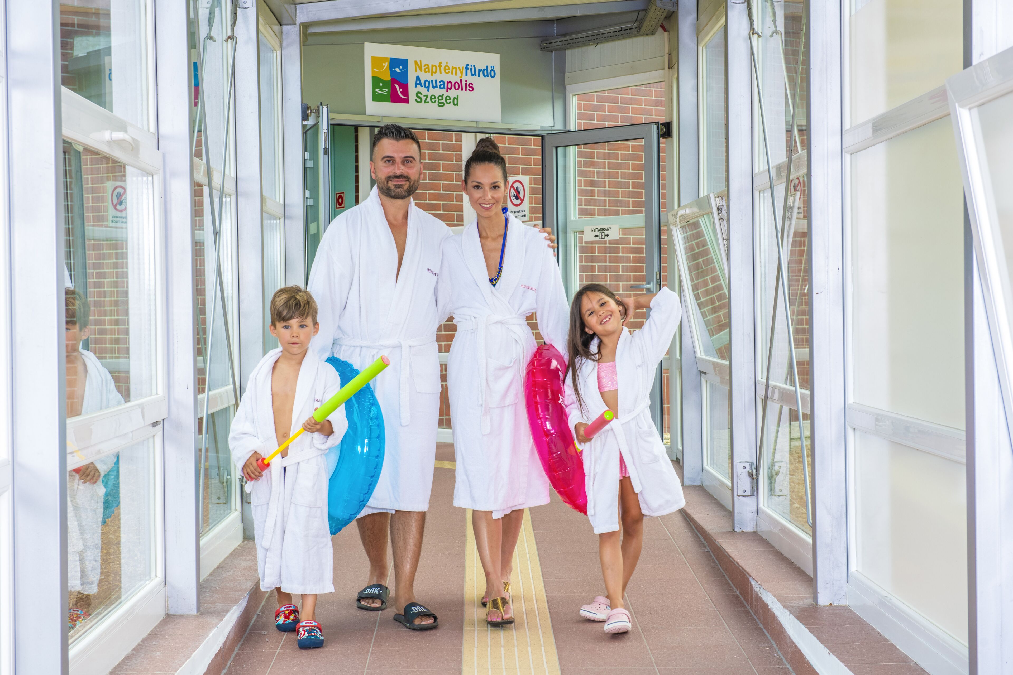 A family in white robes walking through a glass door entrance of a building with a sign reading "Napfényfürdő Aquapolis Szeged".