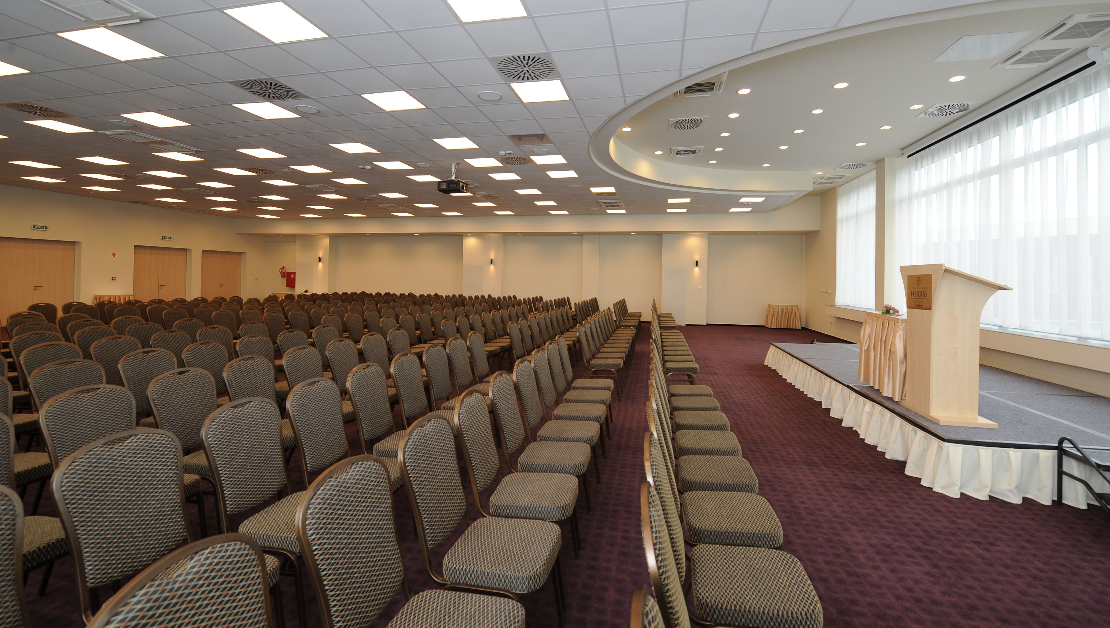 Un salón de conferencias con filas de sillas, un escenario con un atril y ventanas amplias.