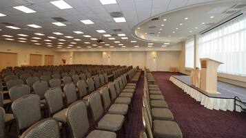 Un salón de conferencias con filas de sillas, un escenario con un atril y ventanas amplias.