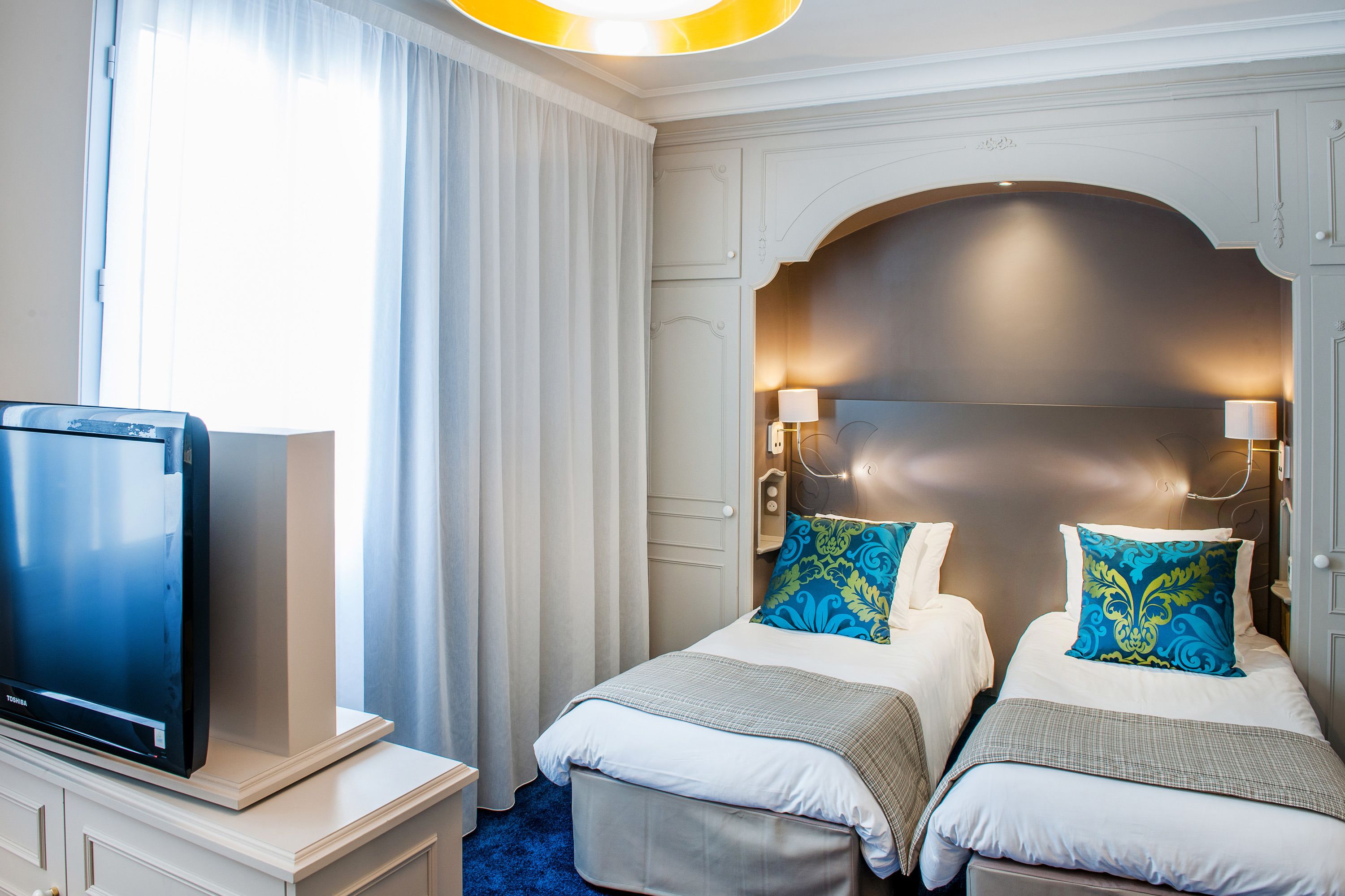 A hotel room with two beds, a TV on a stand, and a large window with curtains.
