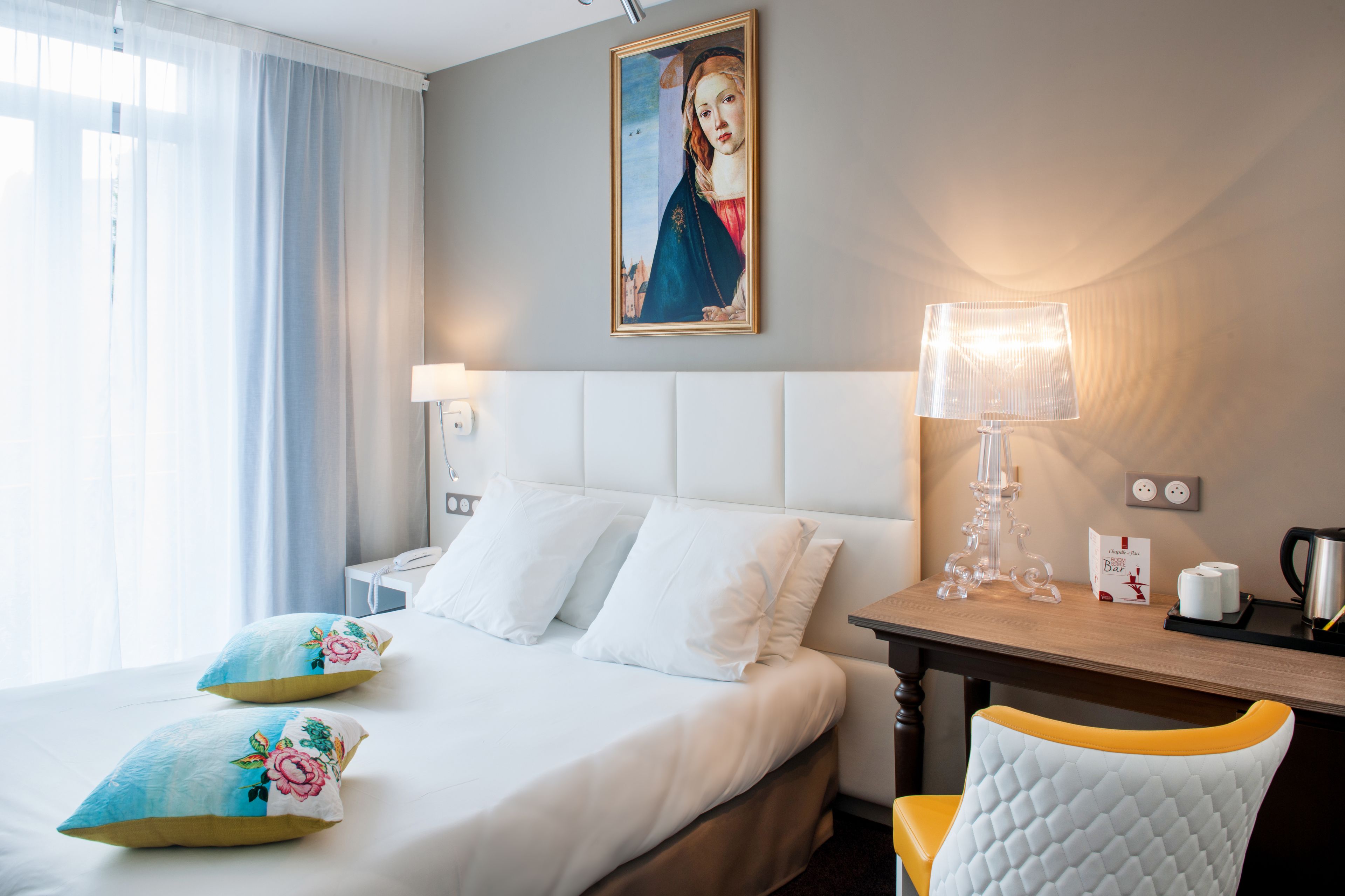 A hotel room with a bed, pillows, a painting, a lamp, a chair, a table, and a coffee maker.