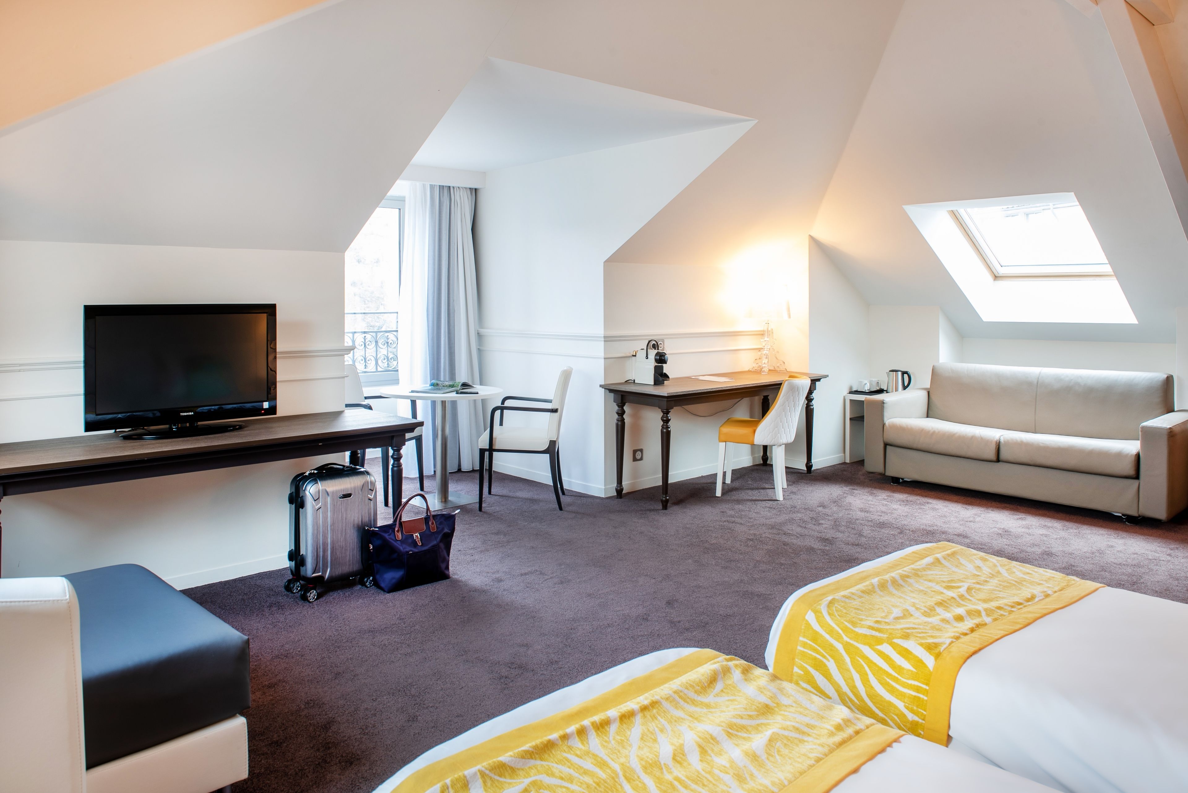 A hotel room with a bed, a desk, a television, a chair, a sofa, and a suitcase.