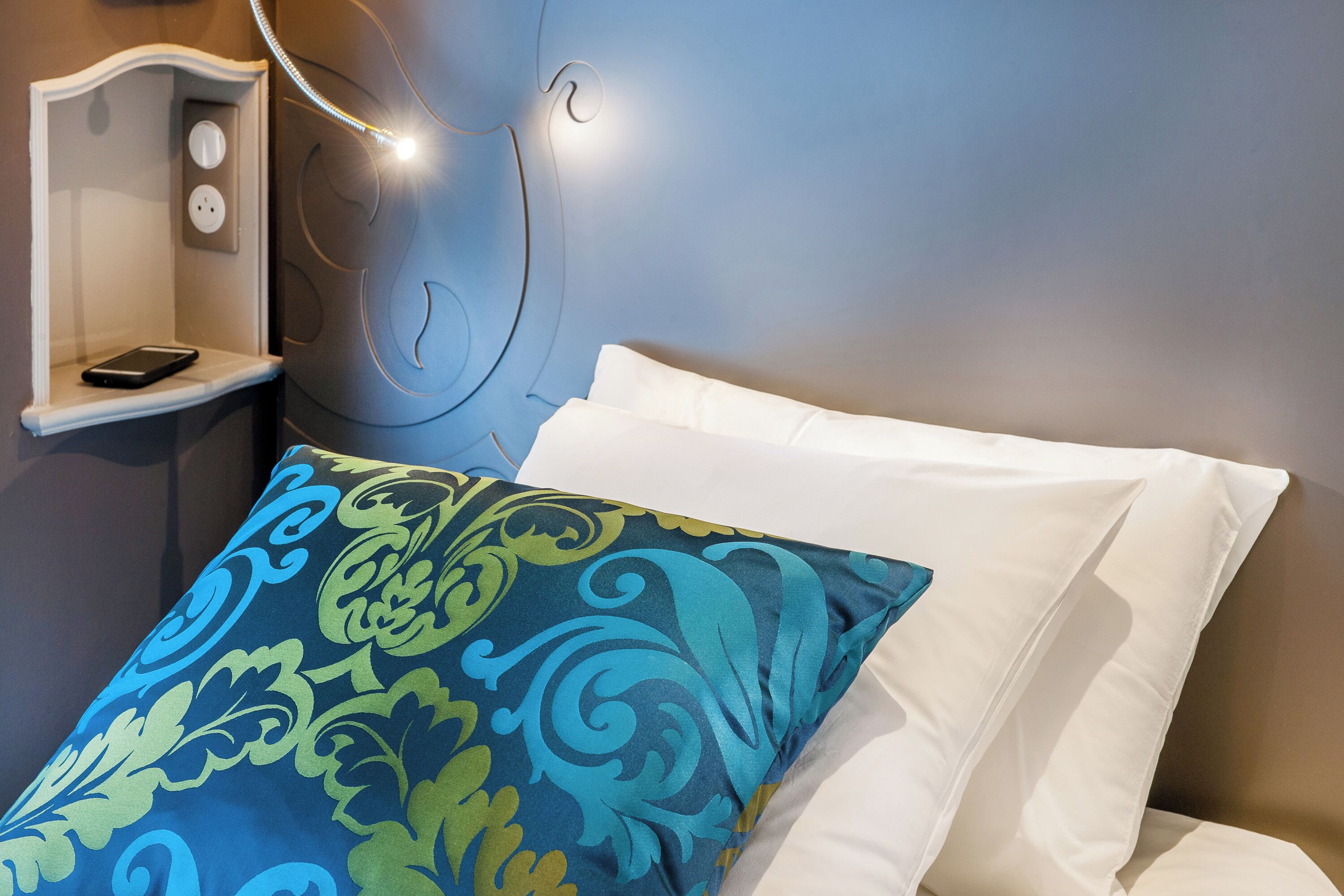 Un letto con un cuscino decorato e un cuscino bianco, un comodino con un telefono e una presa di corrente.