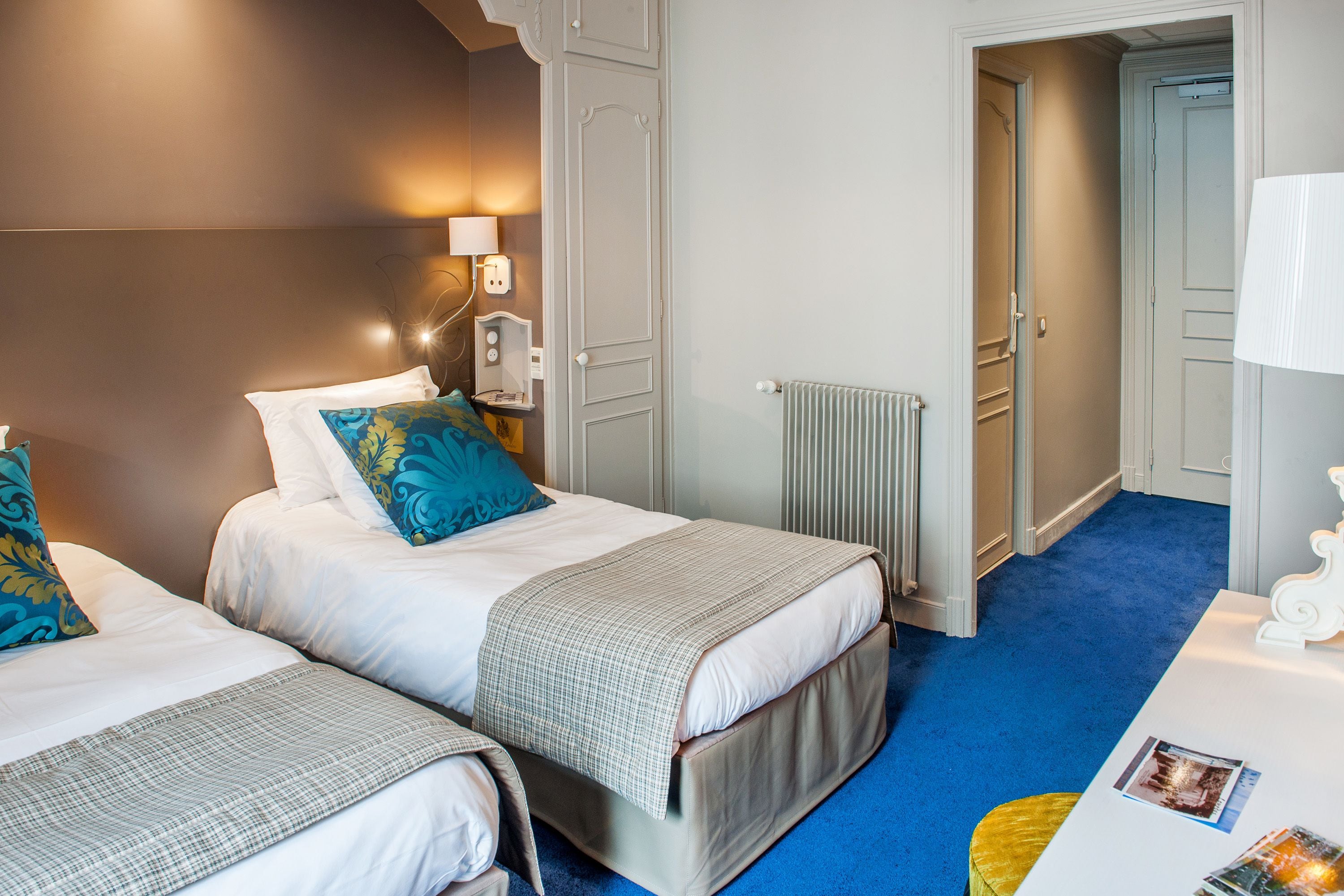 A hotel room with two beds, a radiator, a lamp, and a door leading to another room.