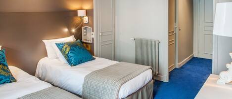 A hotel room with two beds, a radiator, a lamp, and a door leading to another room.