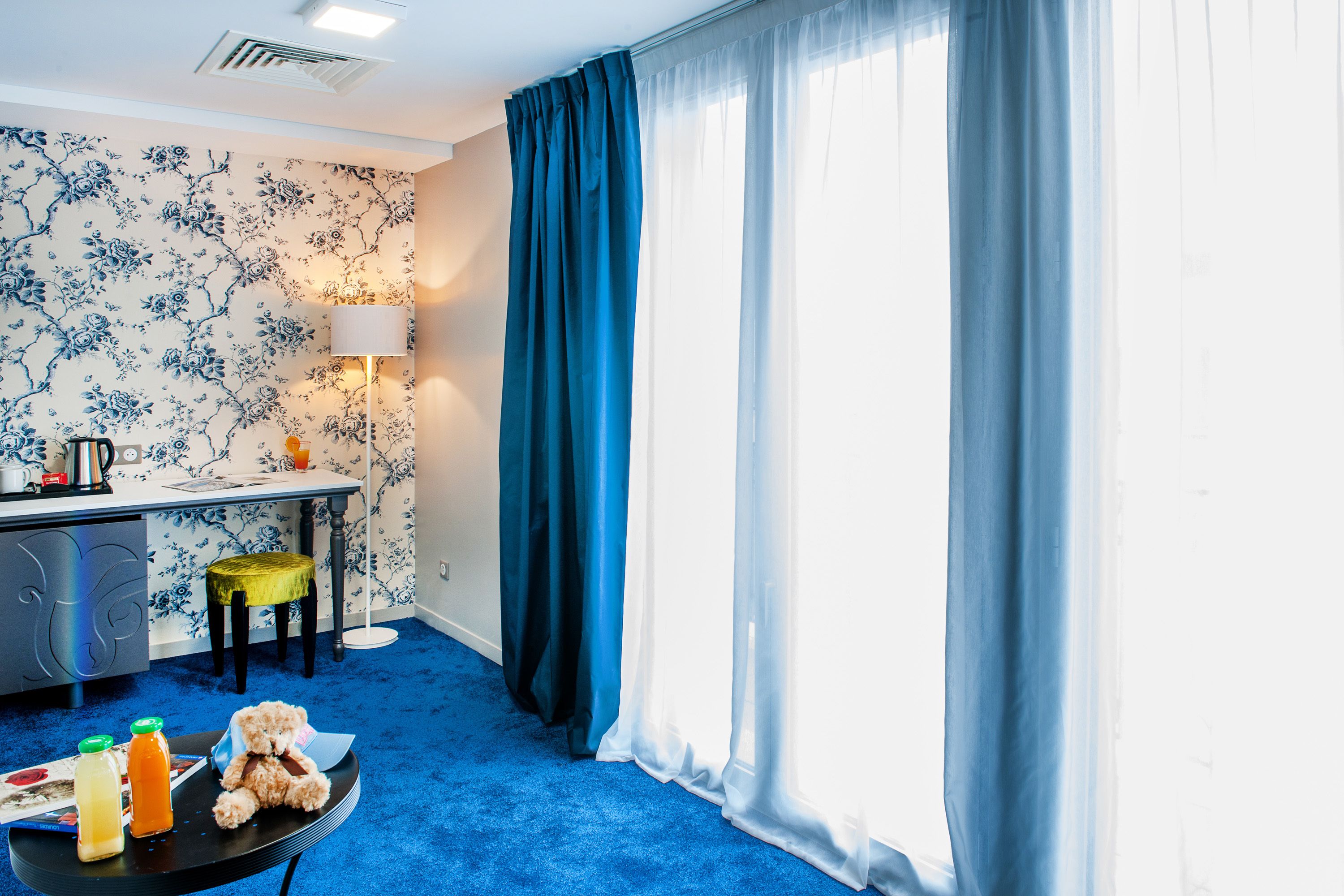 A room with a blue carpet, a desk with a lamp, a chair, a teddy bear, and a window with blue curtains.