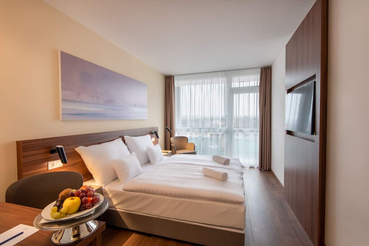 A hotel room with a bed, a desk with a fruit bowl, a TV, and a city view through the window.