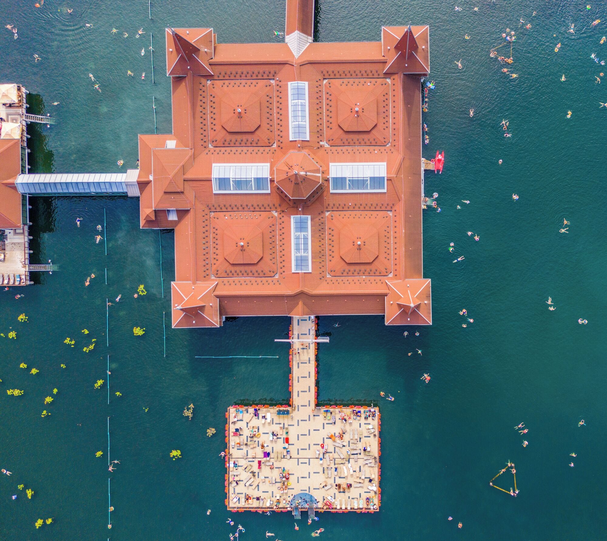 Vista aérea de una estructura flotante cuadrada con un espacio abierto central rodeado de balcones, conectada a un muelle con una zona de piscina.
