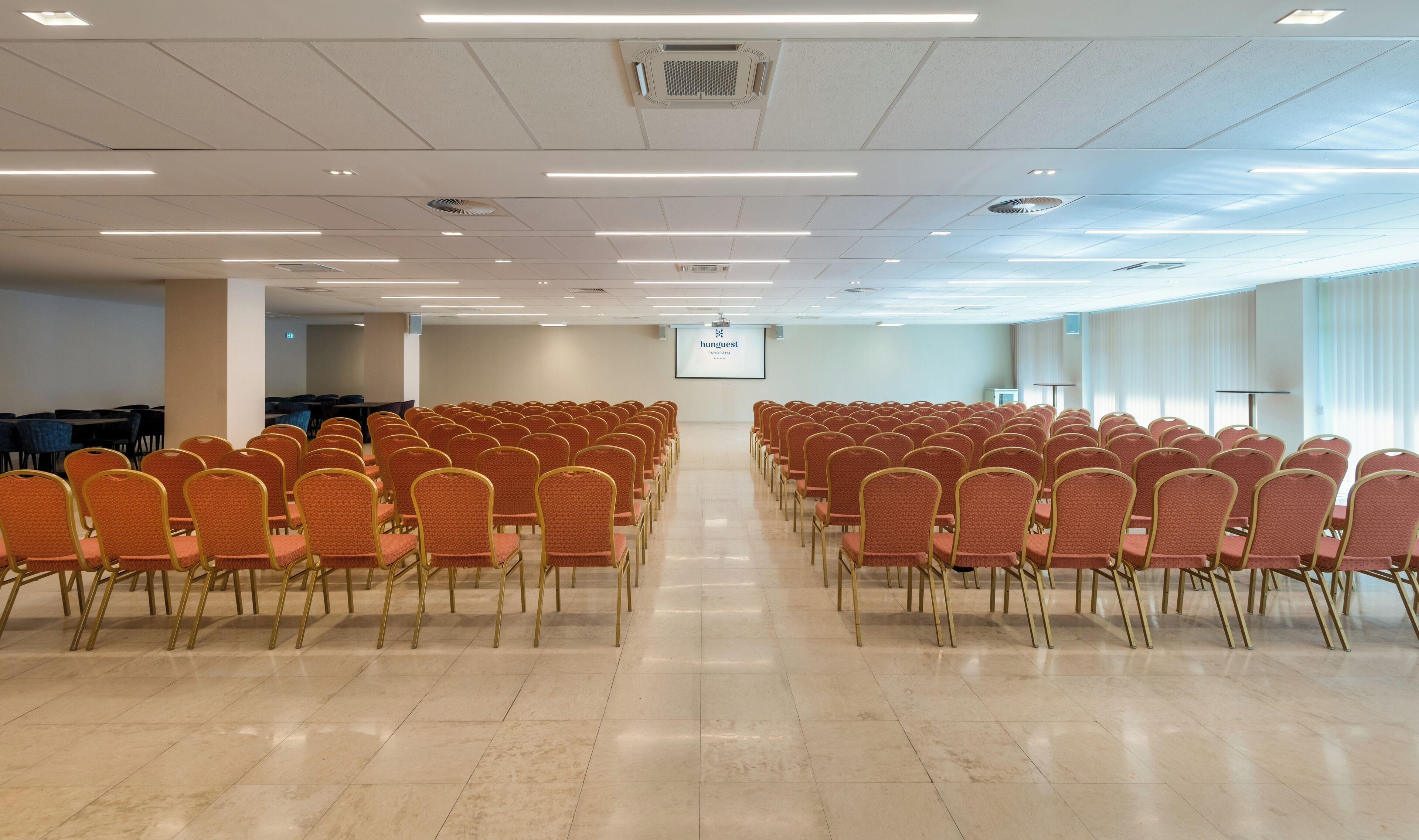 Un salón de conferencias con filas de sillas, una pantalla de proyector y un ventanal grande.