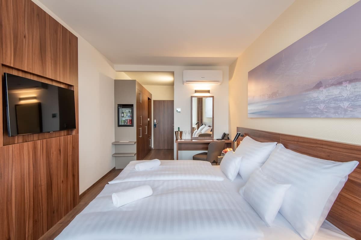 A hotel room with a large bed, a television mounted on a wooden panel, a desk with a chair, and a bathroom visible through an open door.
