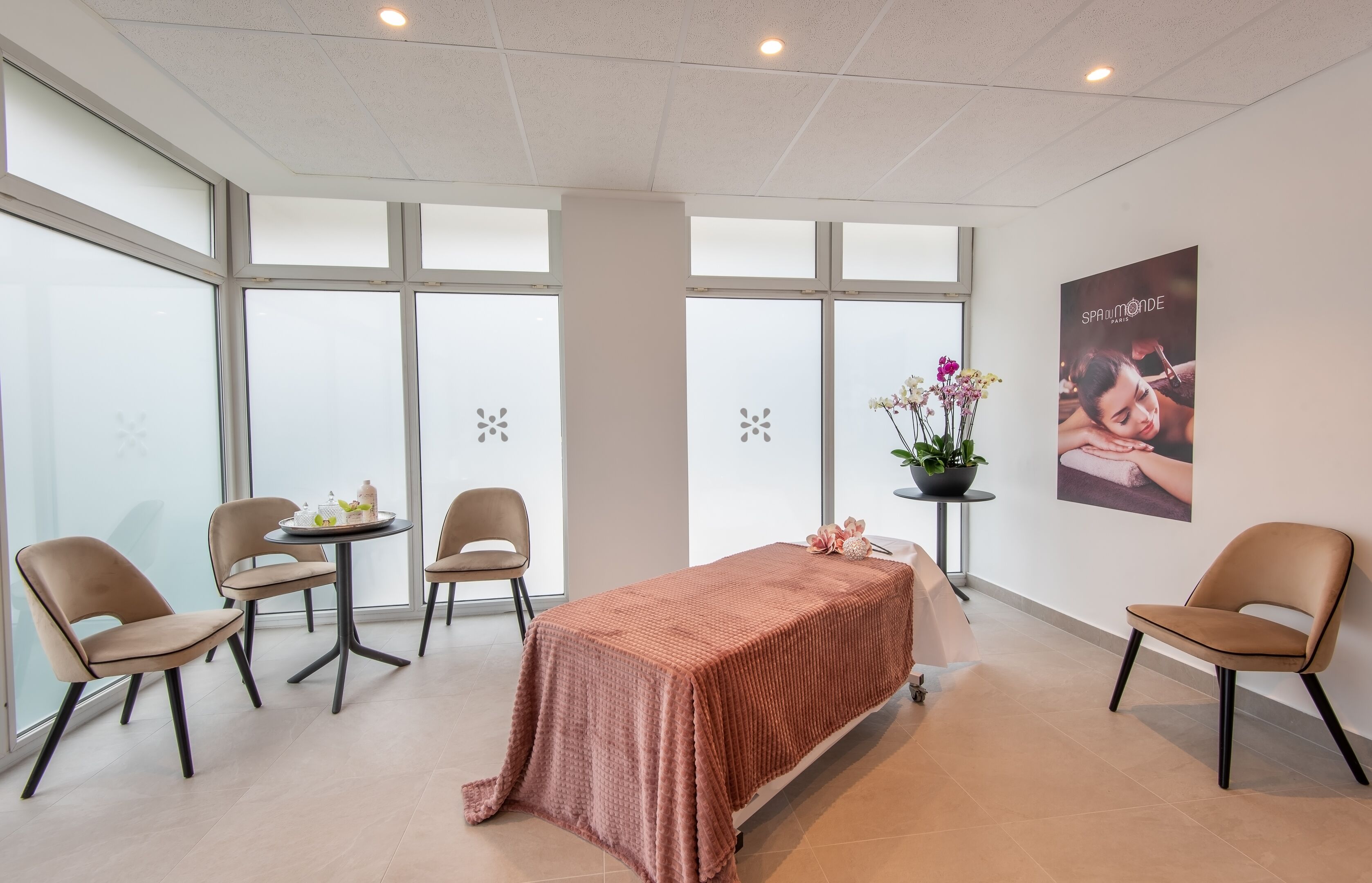 Ett modernt sparum med en massagebänk, två stolar, ett litet bord med en vas med blommor och stora fönster med frostat glas.