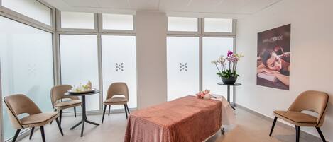 Un spa moderno con una camilla de masaje, dos sillas, una mesita con un florero y ventanas amplias con paneles esmerilados.