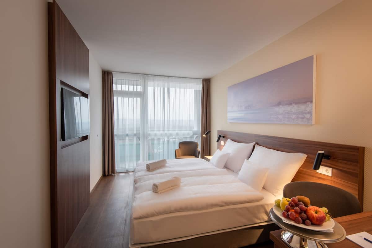 A hotel room with a large bed, a desk with a fruit plate, a chair, a TV, and a balcony with curtains.