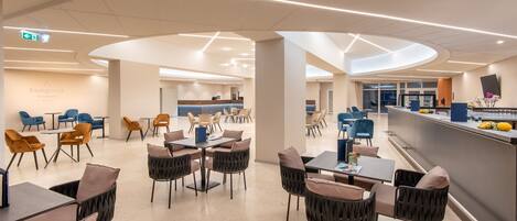 Un hall d’hôtel moderne avec un comptoir d’accueil incurvé, plusieurs espaces de détente avec des chaises et des tables, et une cuisine visible à l’arrière-plan.