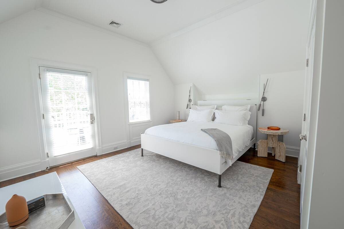 Quarto com cama grande, duas janelas, mesa de cabeceira e um banquinho.