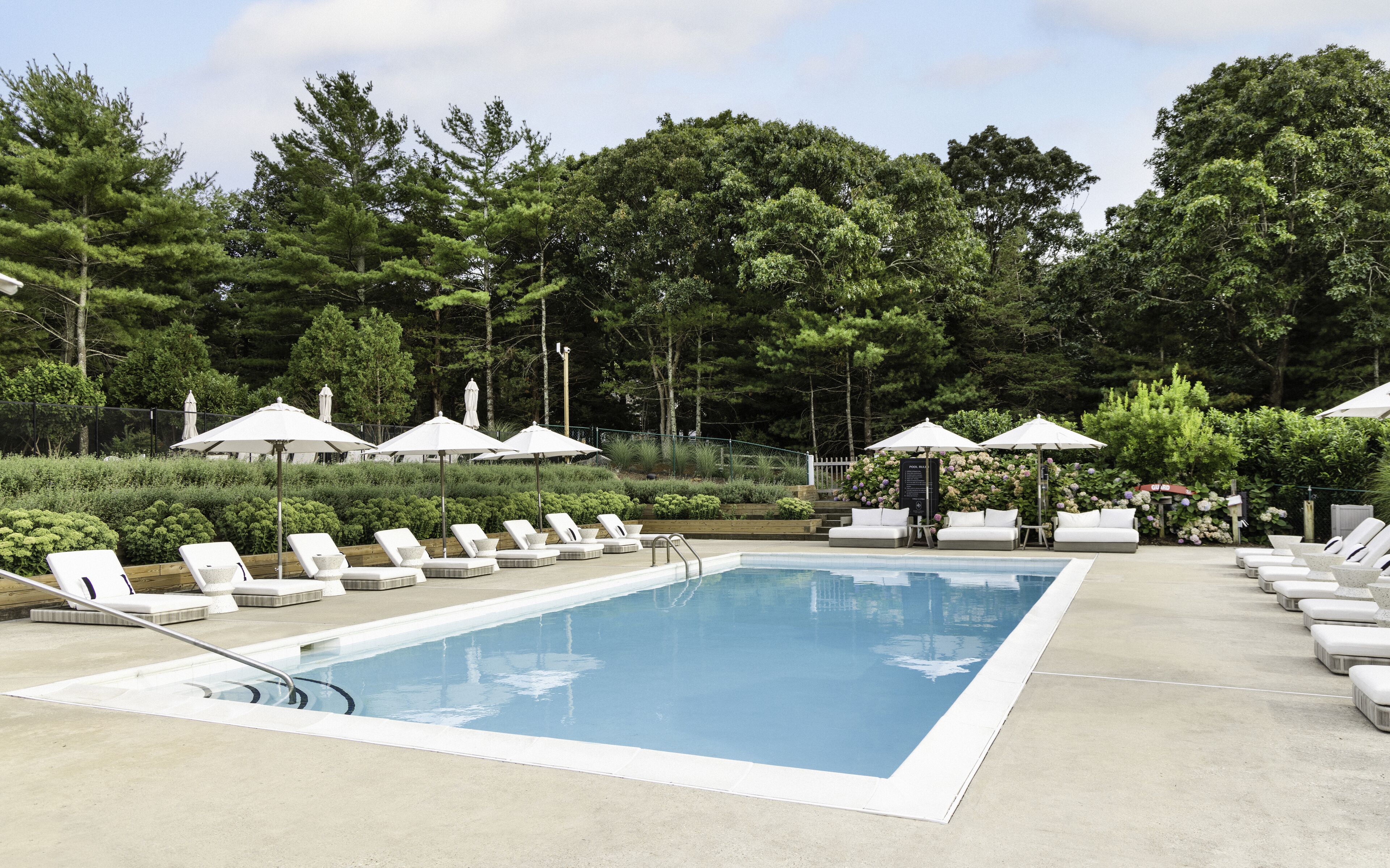 A rectangular pool with lounge chairs and umbrellas, surrounded by trees and greenery.