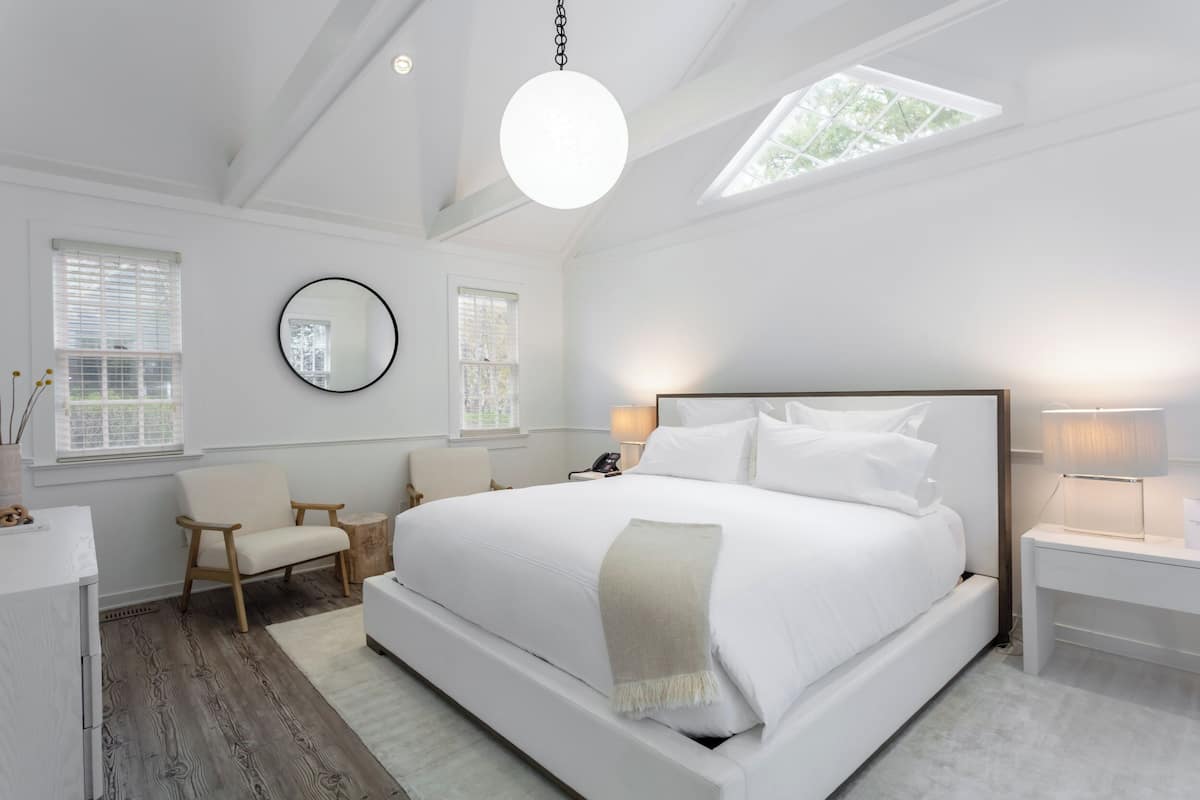 Quarto moderno com cama grande, mesas de cabeceira, espelho redondo e claraboia.