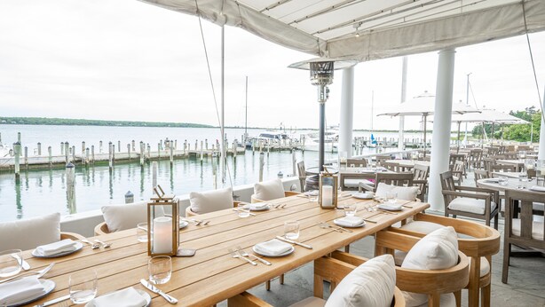 A waterfront restaurant with wooden tables and white chairs, set against a backdrop of a marina and calm water. - EHP Resort & Marina (East Hampton)