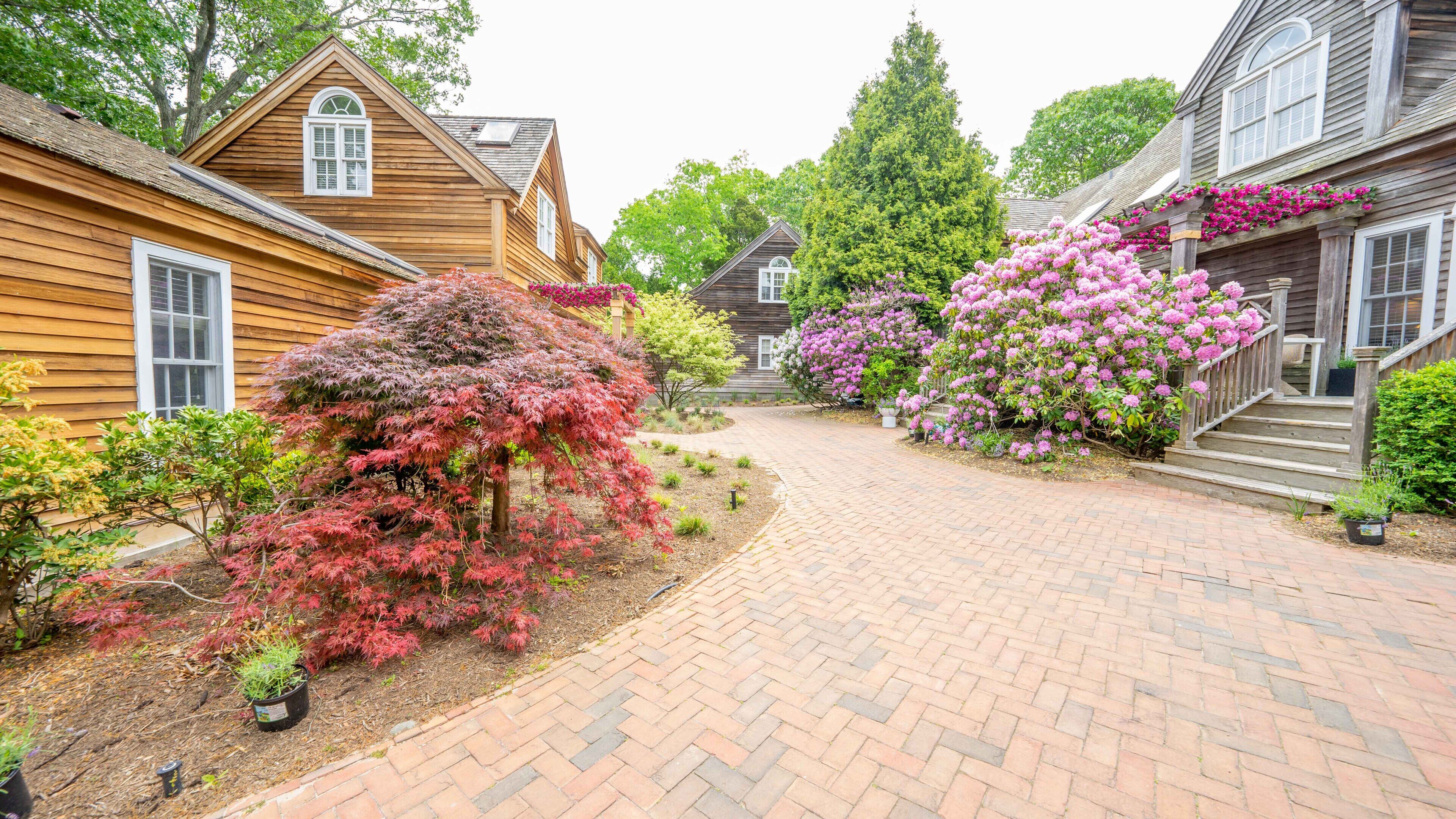 A brick pathway leading to wooden houses with a garden and flowering plants.