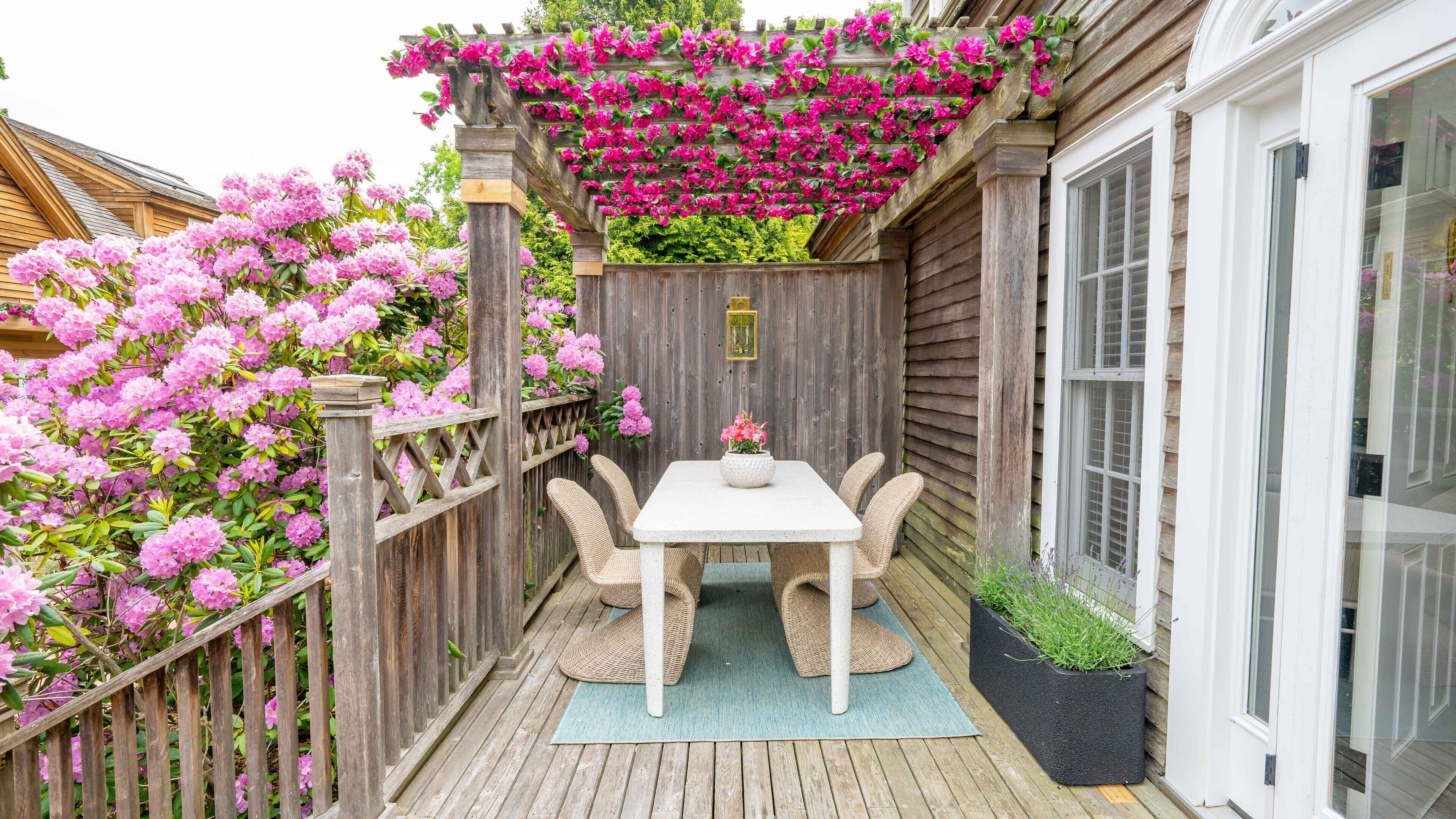 A wooden deck with a table and chairs, surrounded by a wooden fence and blooming pink flowers.