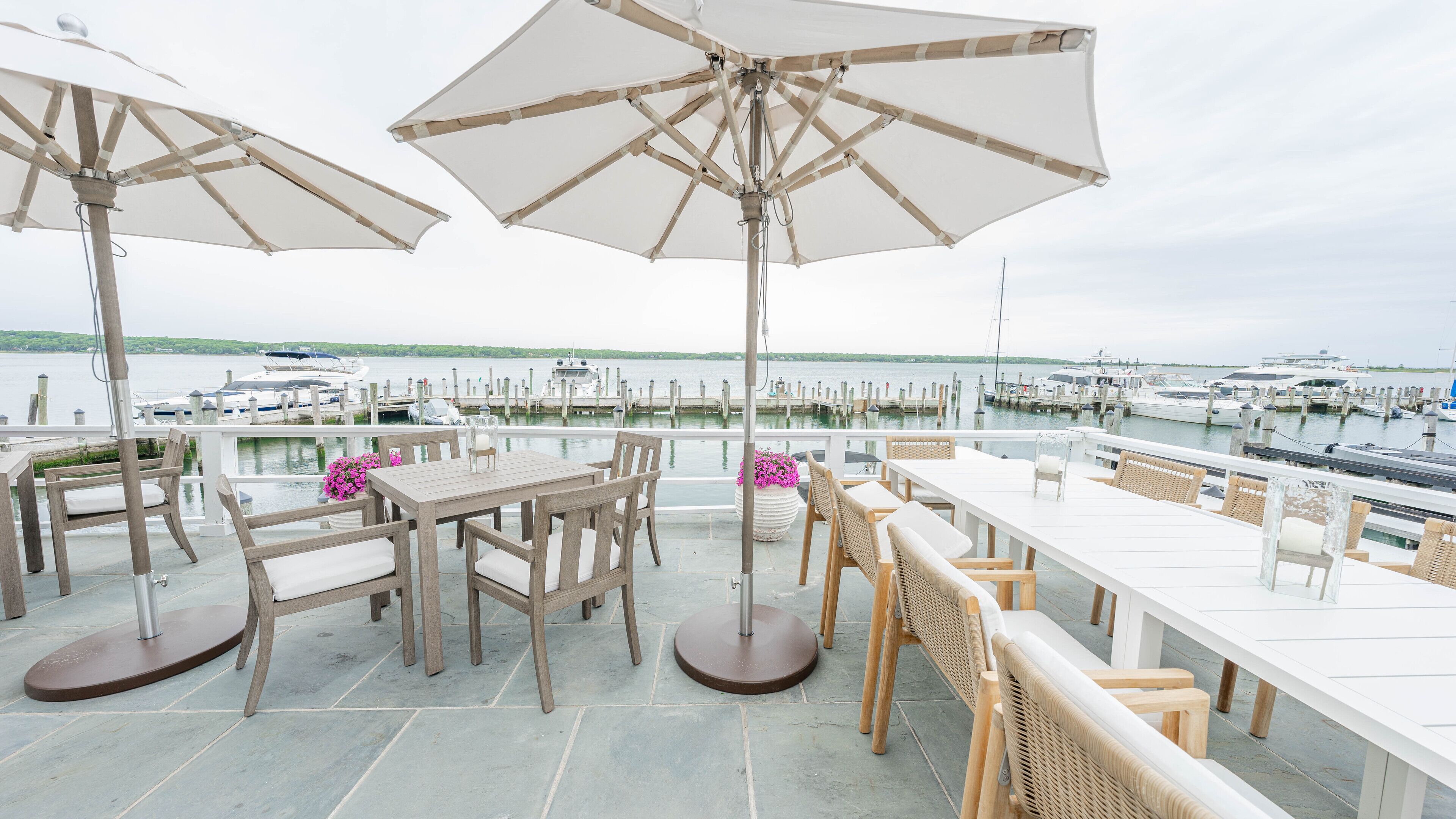 Una terraza con mesas y sillas de madera, grandes sombrillas y vista a un puerto deportivo con barcos.
