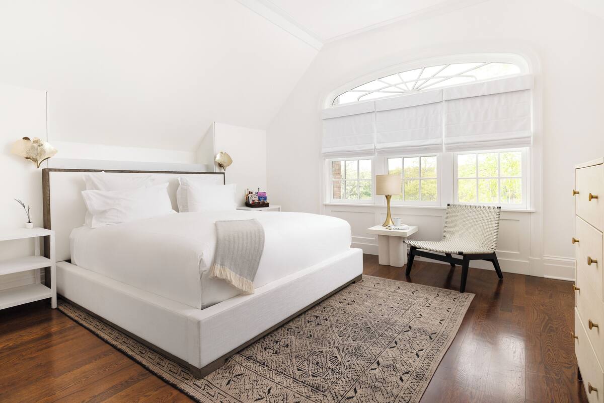 Quarto moderno com uma cama grande, uma mesinha de cabeceira, uma cadeira e uma janela com vista para vegetação.