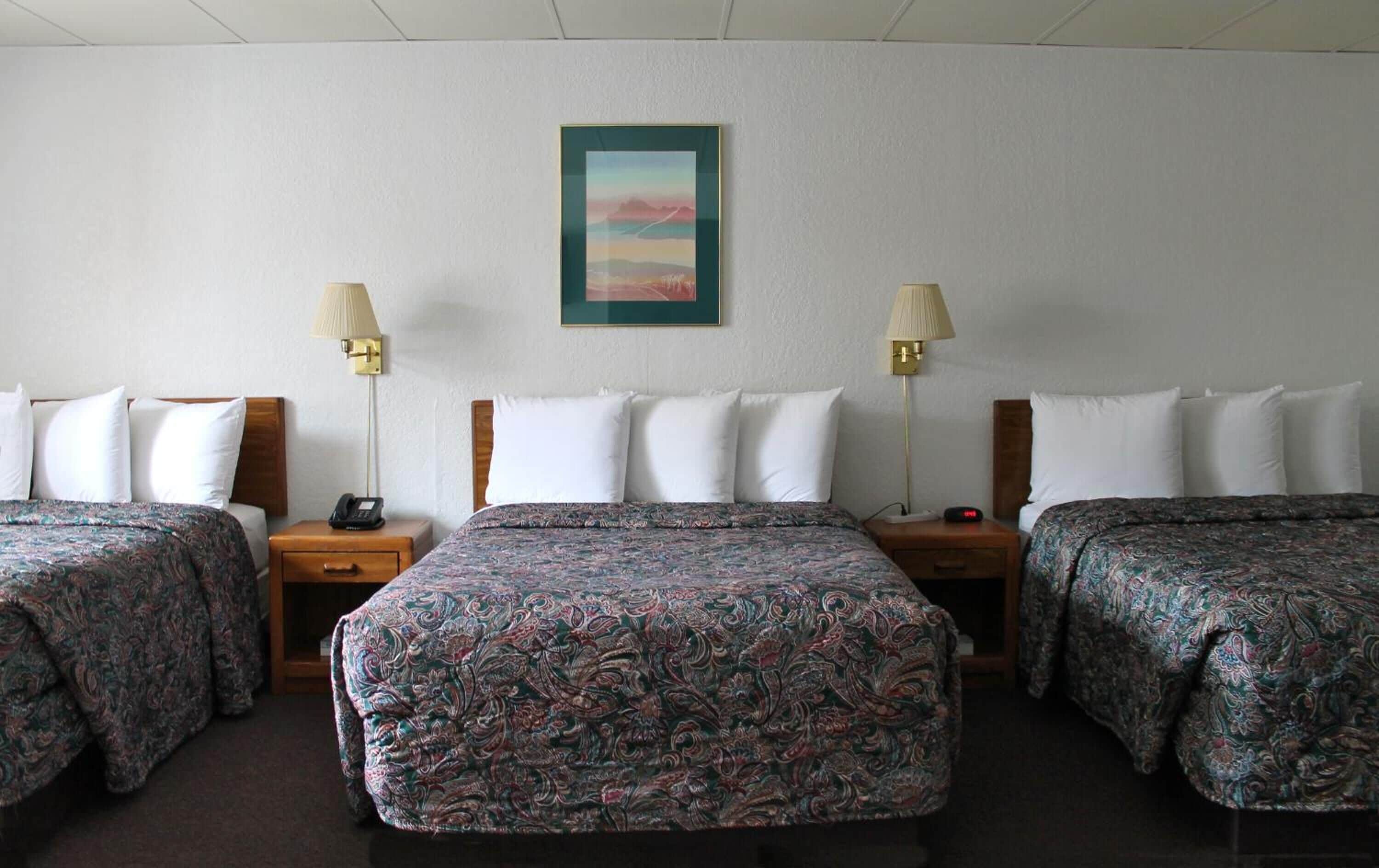 A hotel room with three beds, each with a patterned bedspread, a nightstand with a lamp, and a wall-mounted painting.