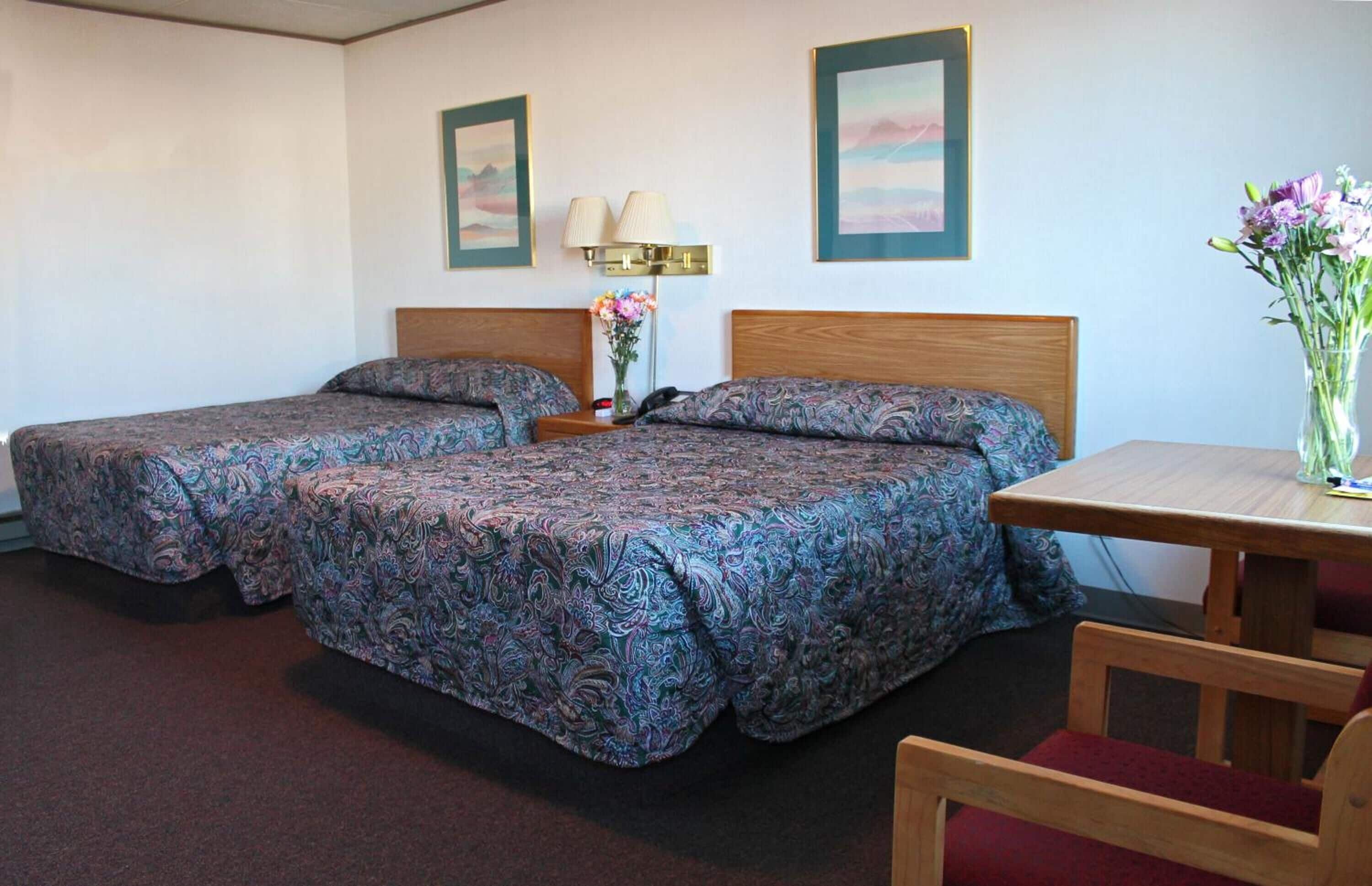 Una camera d'albergo con due letti, una testiera in legno, un comodino con una lampada, un vaso con fiori e un tavolino con una sedia.