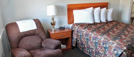 A hotel room with a brown leather chair, a wooden nightstand with a lamp, a bed with a patterned bedspread, and a wooden headboard.