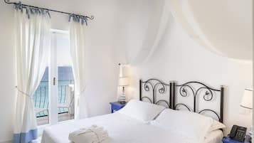 A hotel room with a bed, a nightstand with a lamp, a balcony with a chair, and a view of the sea.