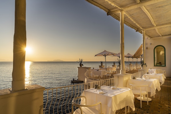 A seaside restaurant with outdoor seating overlooking the sea, featuring white tables and umbrellas.
