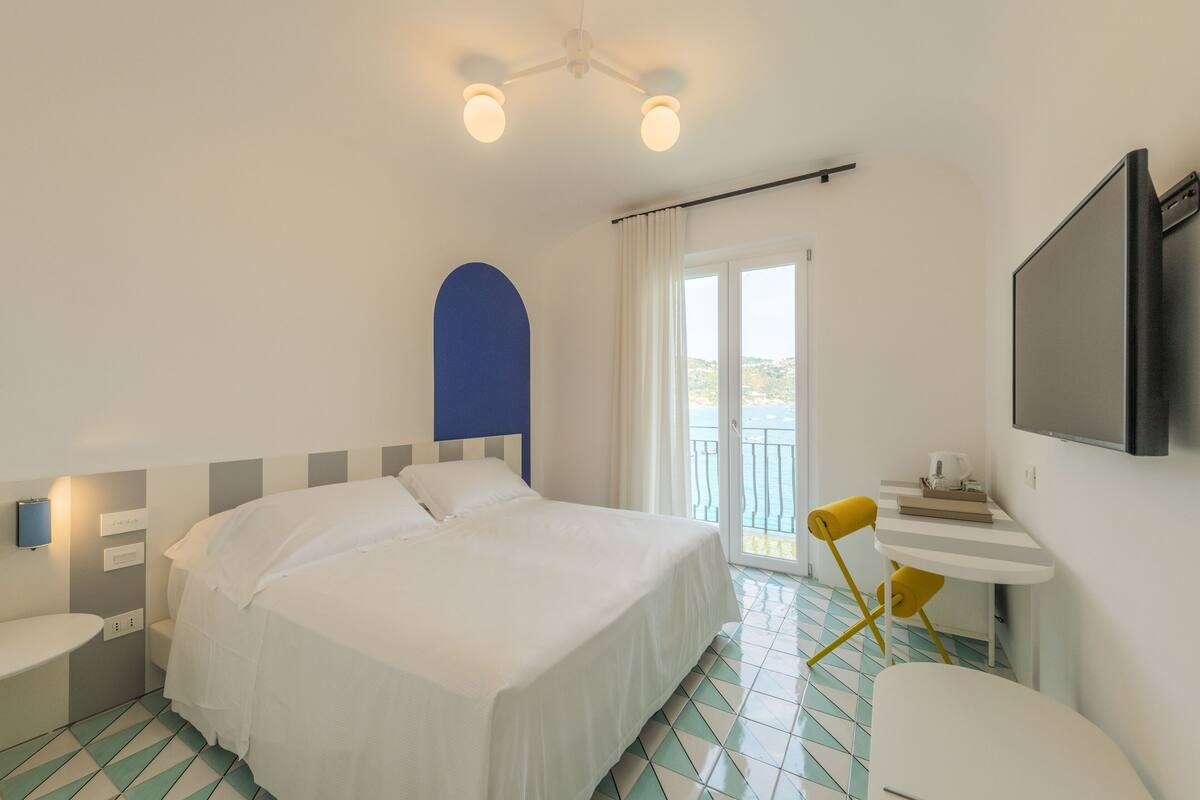 Habitación de hotel con cama, una mesita con silla, televisión y balcón con vistas.