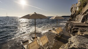 Une plage avec des parasols et des chaises longues, une falaise rocheuse et un voilier à l’horizon.