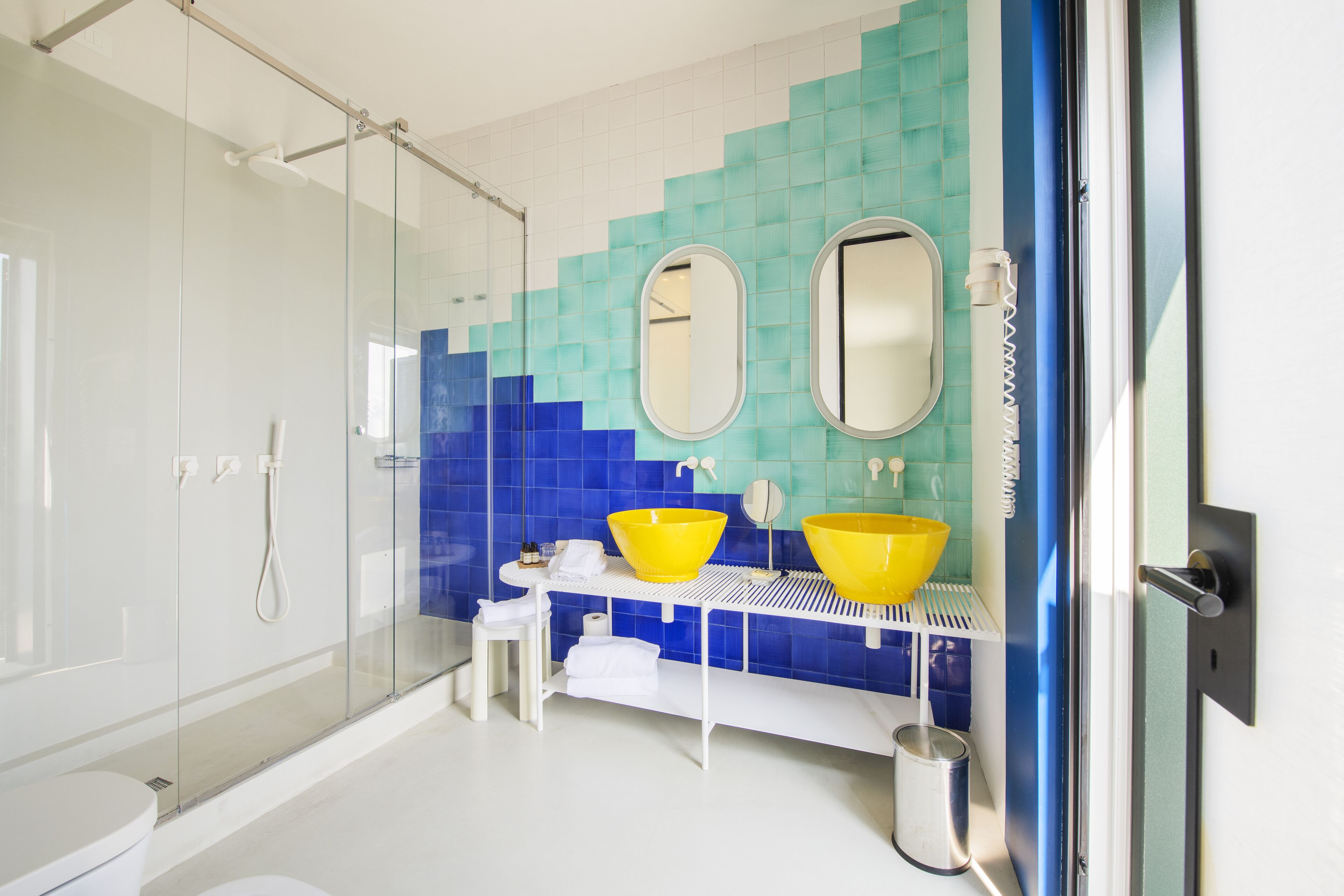 Een moderne badkamer met een douchecabine van glas, twee gele wastafels en een wand betegeld met blauwe tegels.