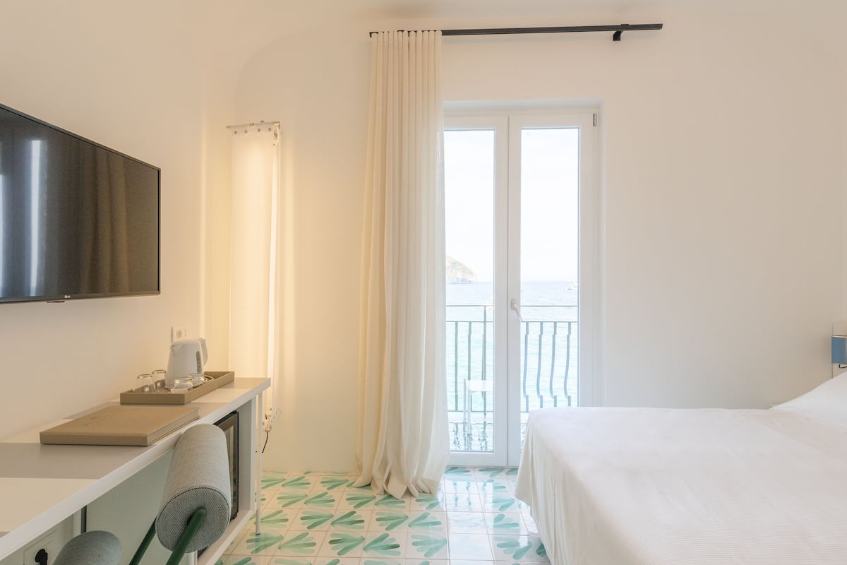 Habitación de hotel con cama, televisión, escritorio con hervidor y vista al mar desde el balcón.