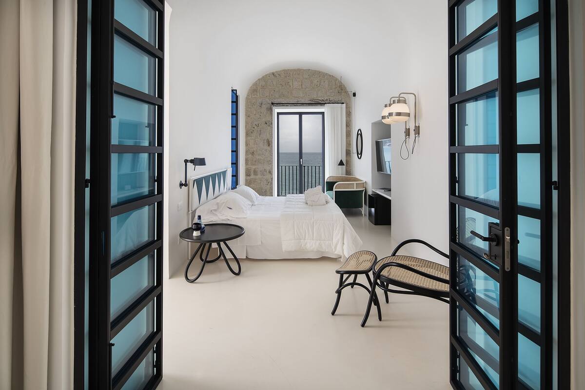 Un dormitorio con cama, una mesita, dos sillas, televisor y puerta al balcón.