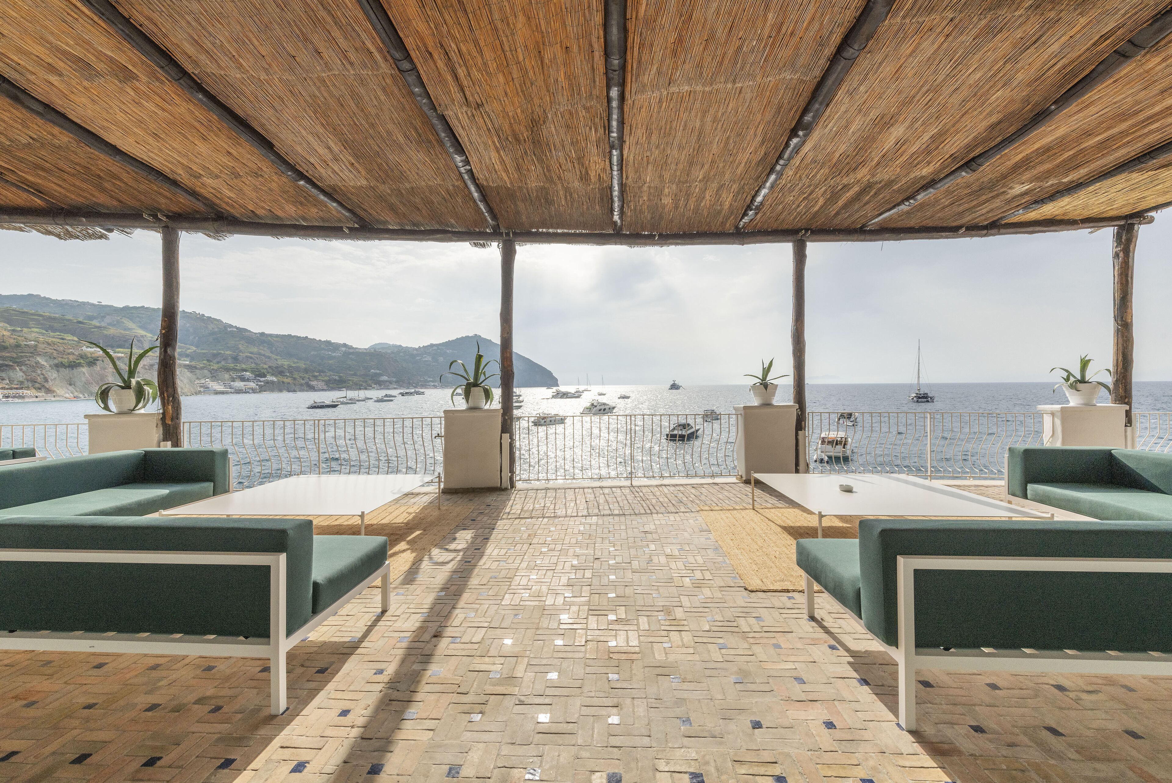 An outdoor seating area with a view of the sea, featuring a thatched roof, green cushioned seating, and a tiled floor.