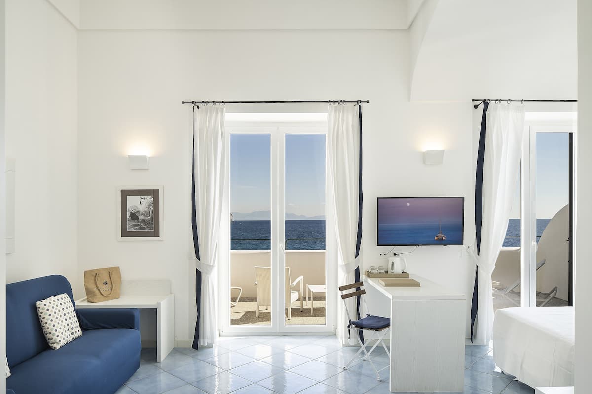 Una habitación con sofá, escritorio, silla y cama. Hay un televisor en la pared y una ventana con cortinas.
