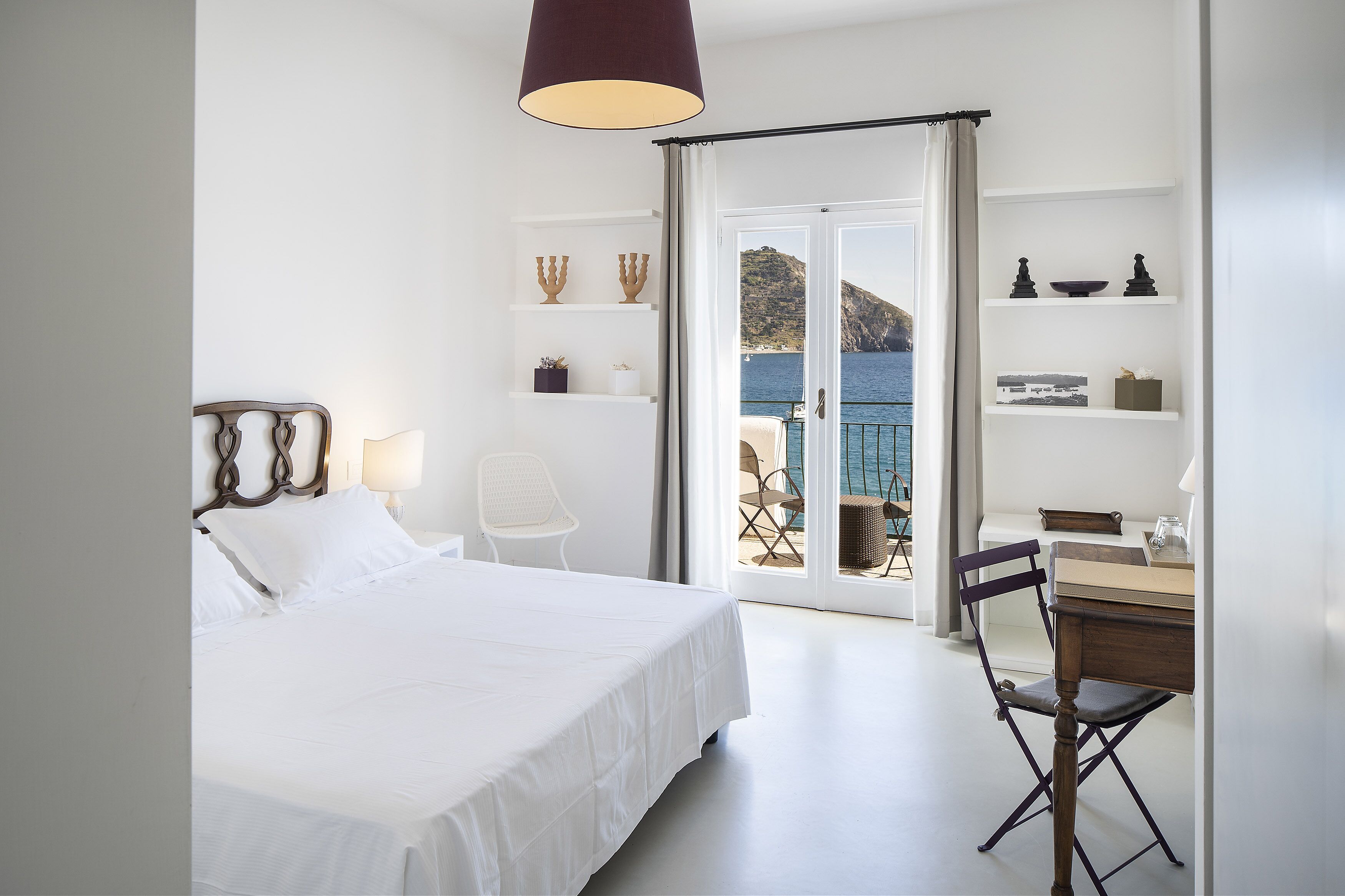 A bedroom with a bed, a desk, and a chair. There is a view of the sea through the window.