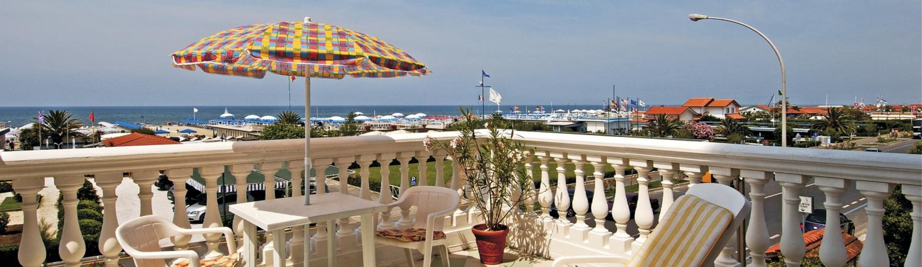 Ein Balkon mit weißen Stühlen und einem bunten Sonnenschirm mit Blick auf eine Marina voller Boote und Häuser.