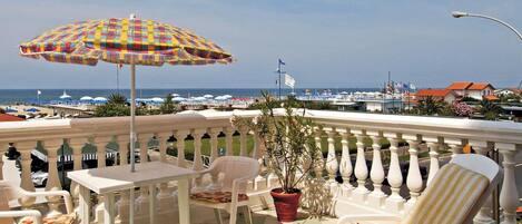 Ein Balkon mit weißen Stühlen und einem bunten Sonnenschirm mit Blick auf eine Marina voller Boote und Häuser.