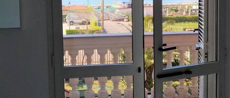 A room with a bed, a view through a sliding glass door to an outdoor area with a tiled floor, a railing, and a clear sky.