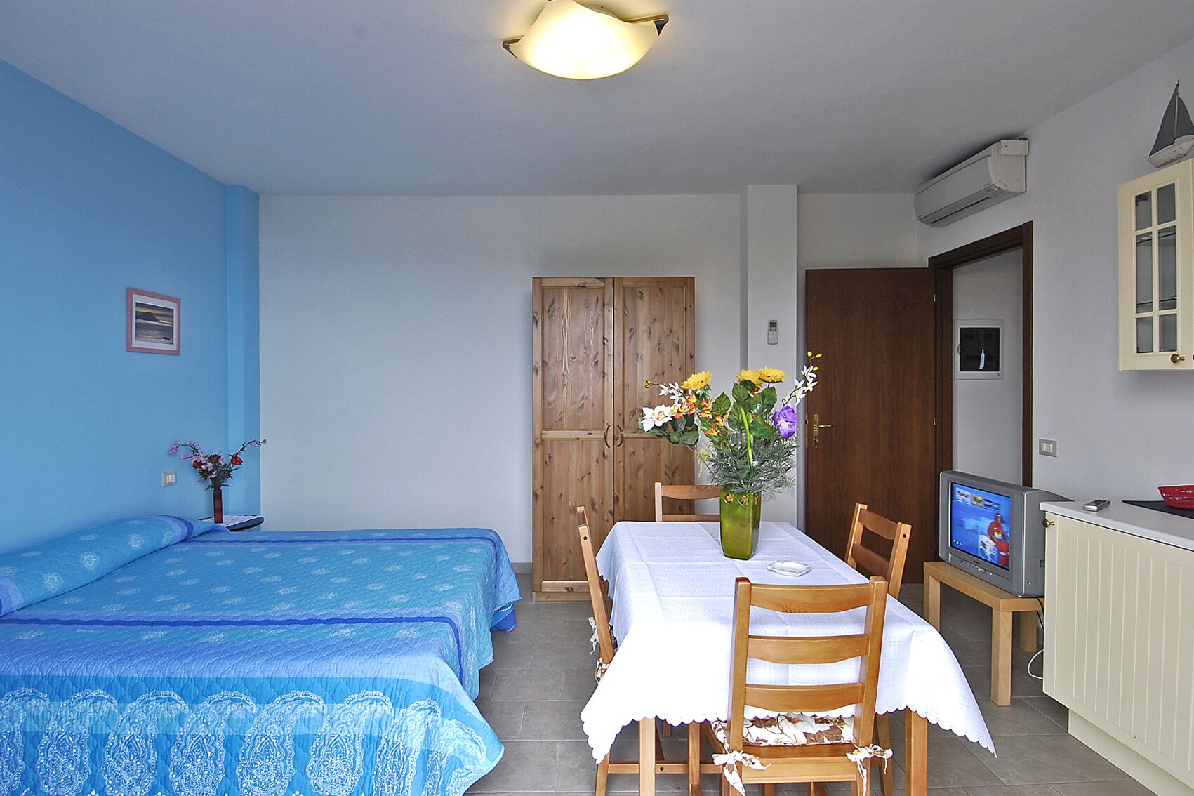 A room with two beds, a dining table with chairs, a vase with flowers, a television, and an air conditioning unit.
